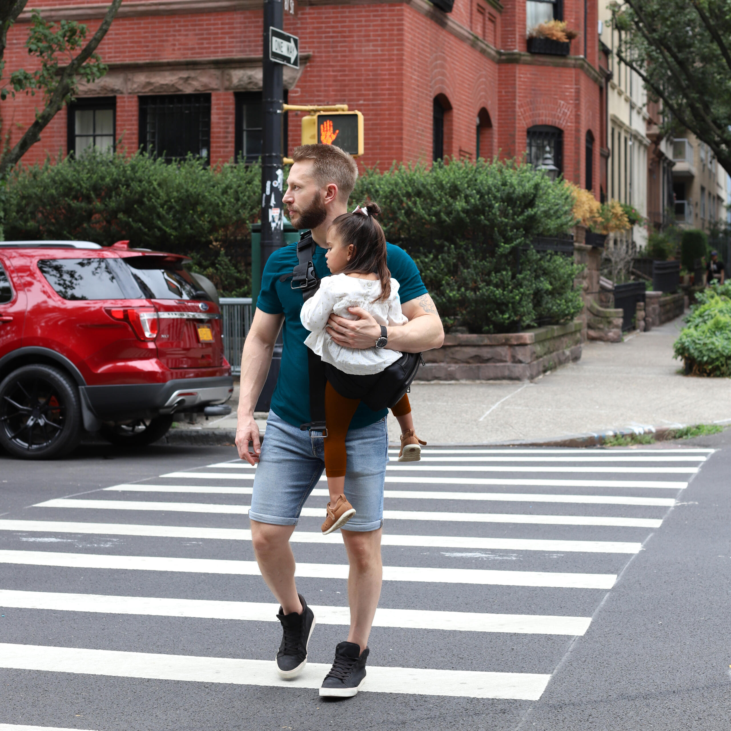 Dad carrying a toddler in a sleek black Jacana convertible baby carrier and diaper bag while crossing a city street.