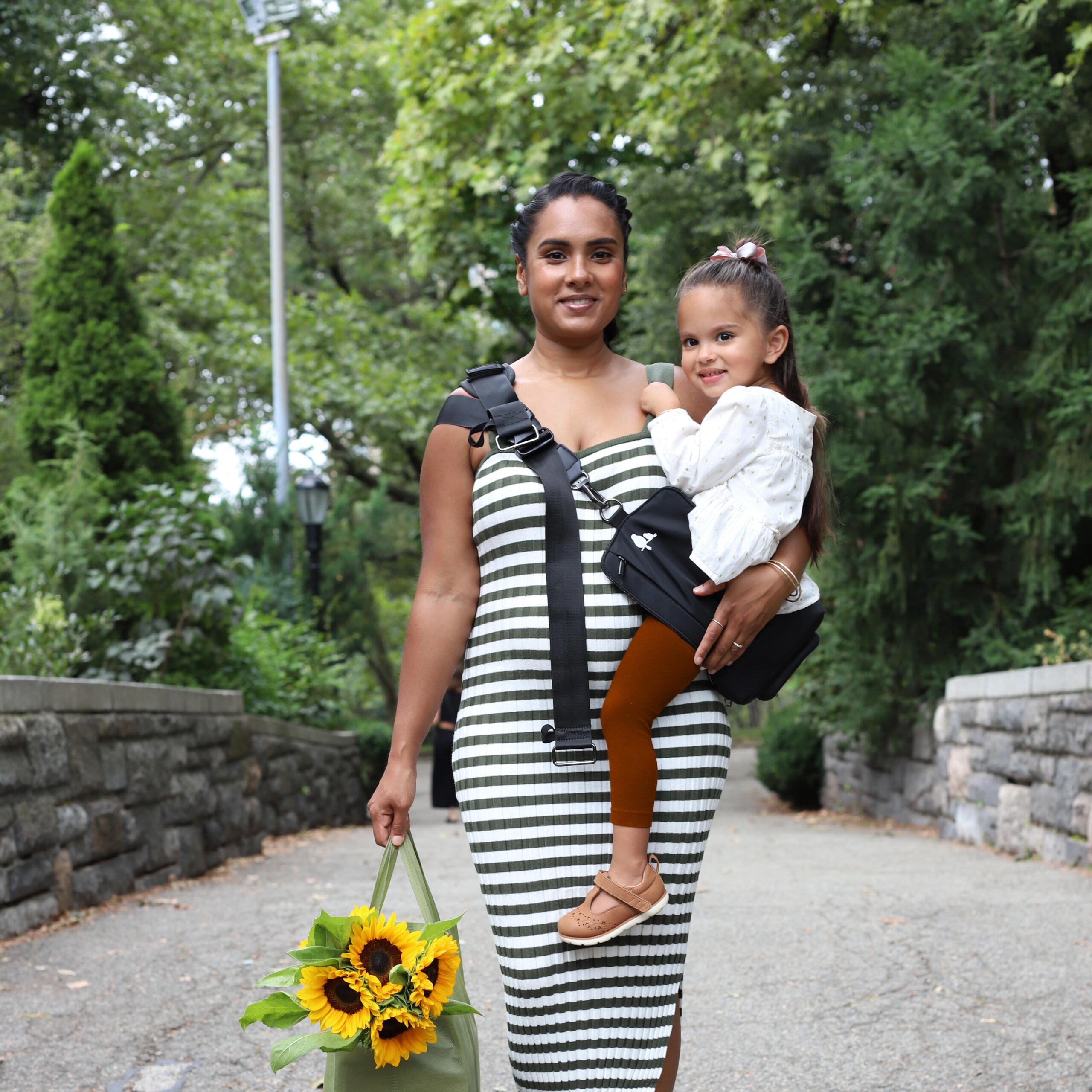 Mom carrying a toddler in a sleek black Jacana convertible baby carrier and diaper bag while walking through a city park.